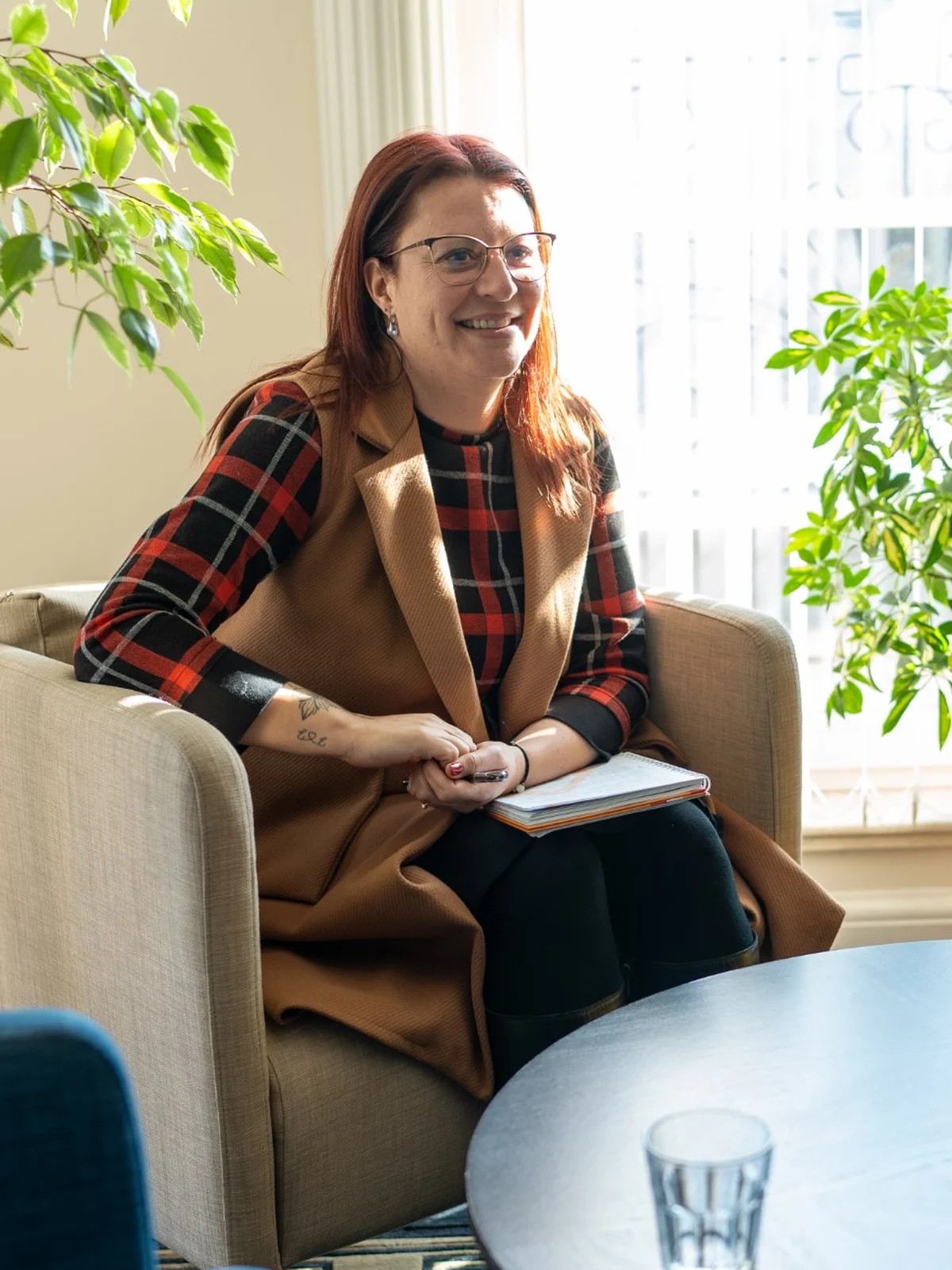 Smiling woman with glasses sits in a beige armchair holding a notebook, surrounded by green plants.