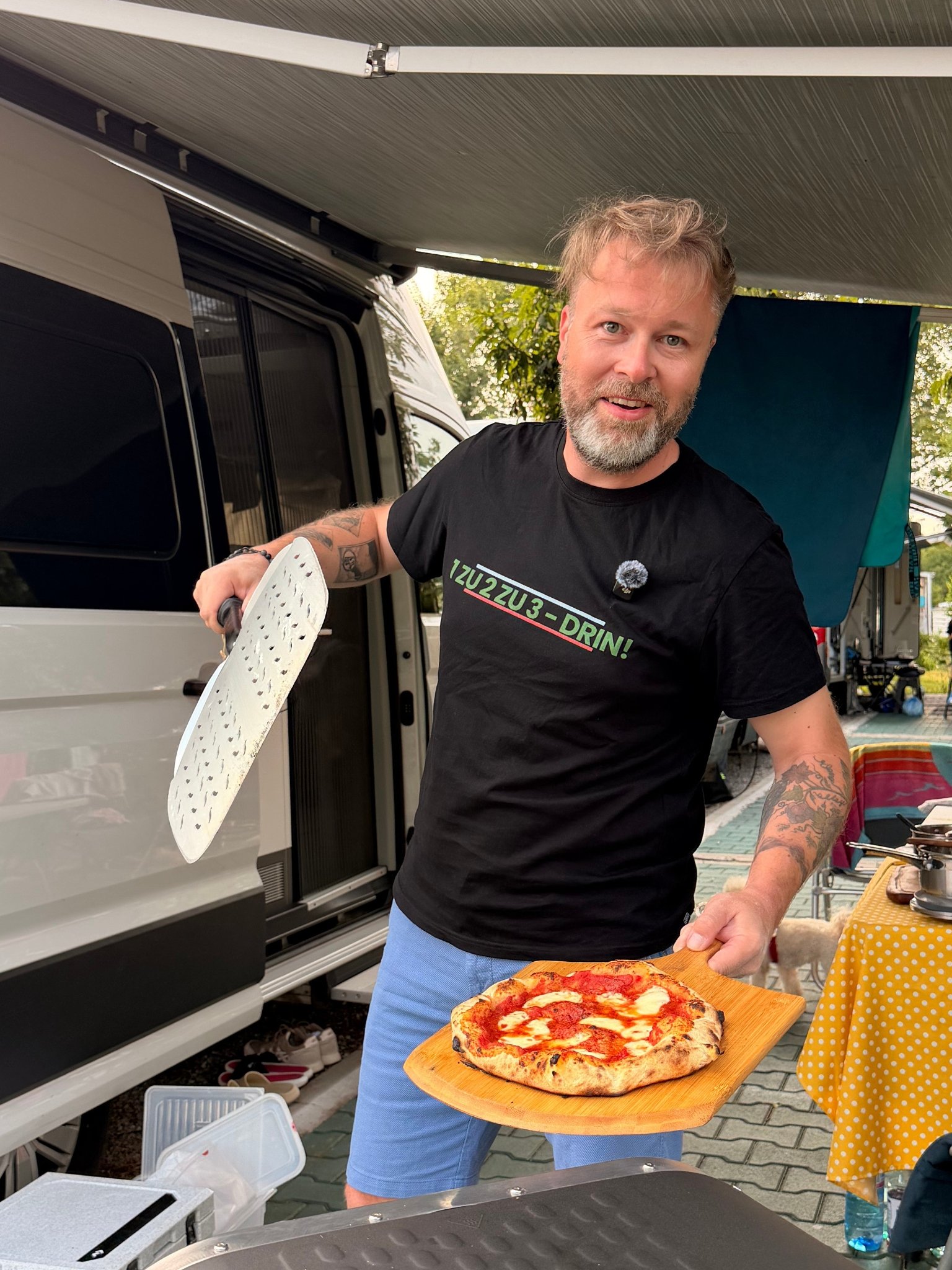 Bearded man holds a pizza and peel beside a white camper van.